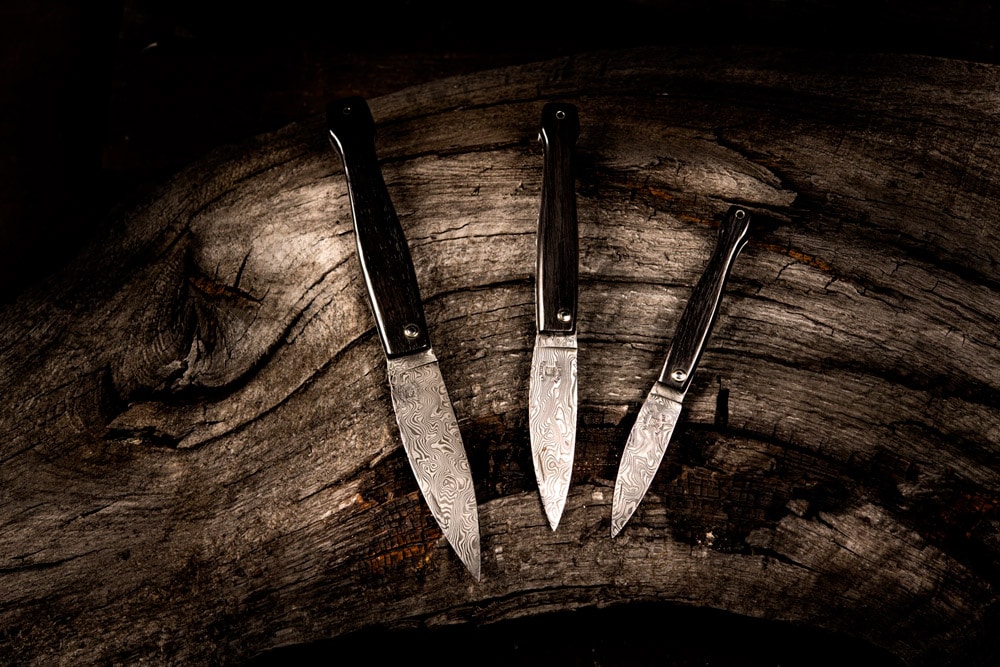 Three pocket knives with intricately patterned damascus blades and dark handles, fanned out on an aged wooden stump.