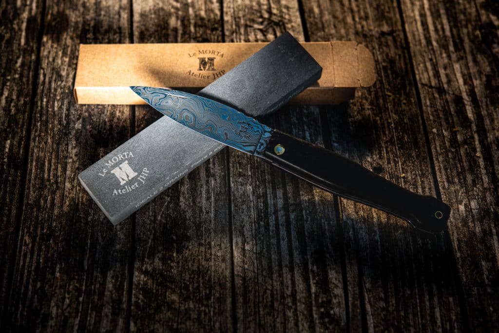 A folding knife with a black and blue Damascus steel blade, matte black handle and complementary whetstone, presented alongside its rustic packaging box.