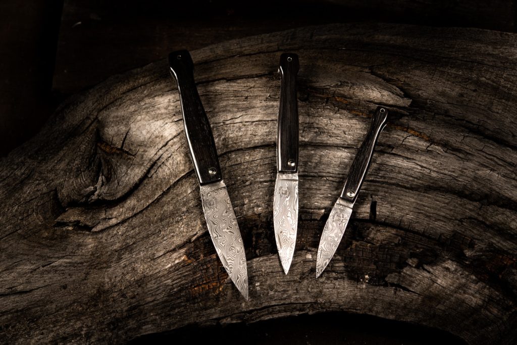 Trois couteaux de poche avec des lames en damas aux motifs complexes et des manches sombres, disposés en éventail sur une souche en bois vieilli.