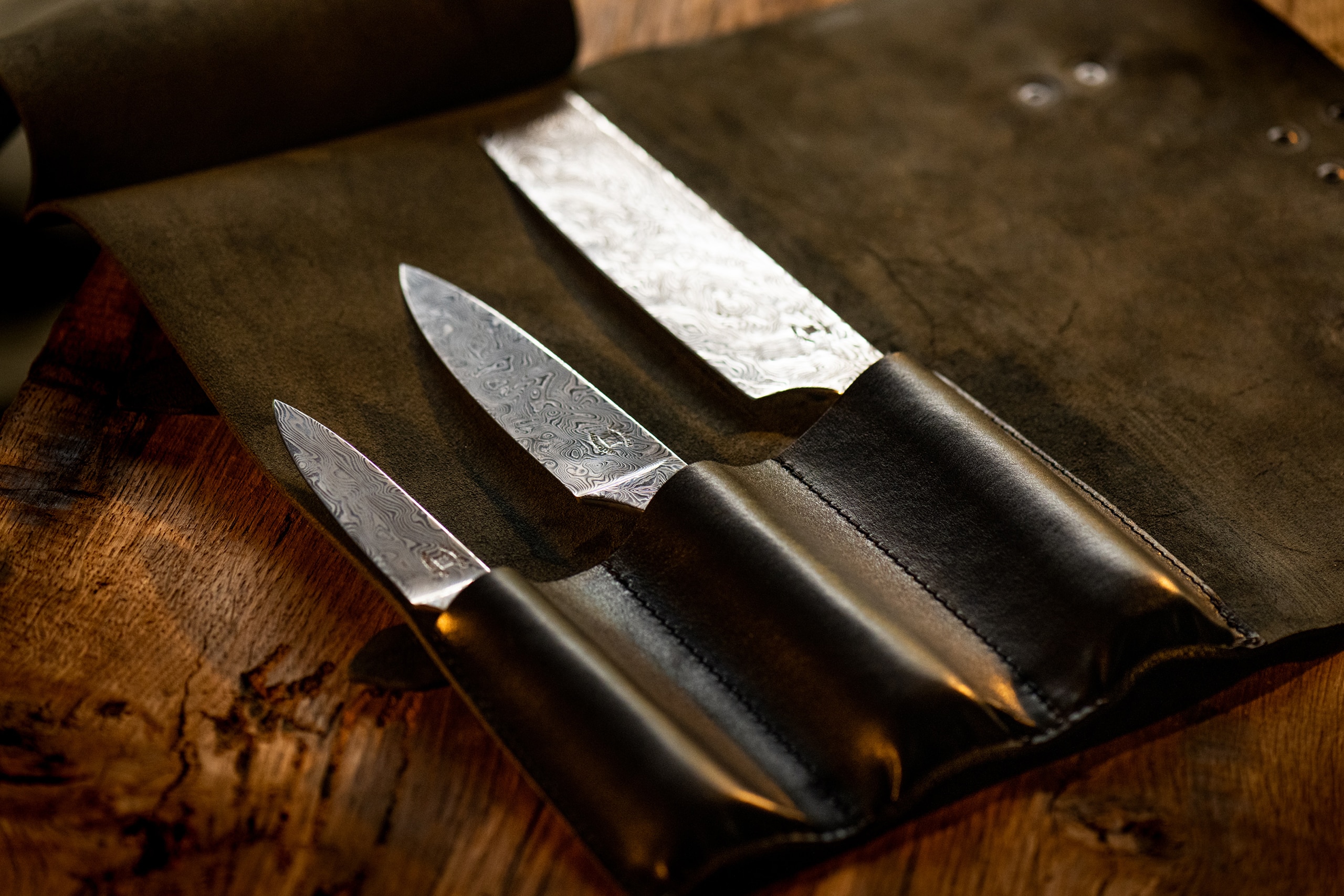 Trois couteaux avec lames en acier damassé, exposés ouverts sur un étui en cuir brun foncé, posés sur une surface en bois vieilli, évoquant un ensemble de couteaux d'artisanat français de qualité.