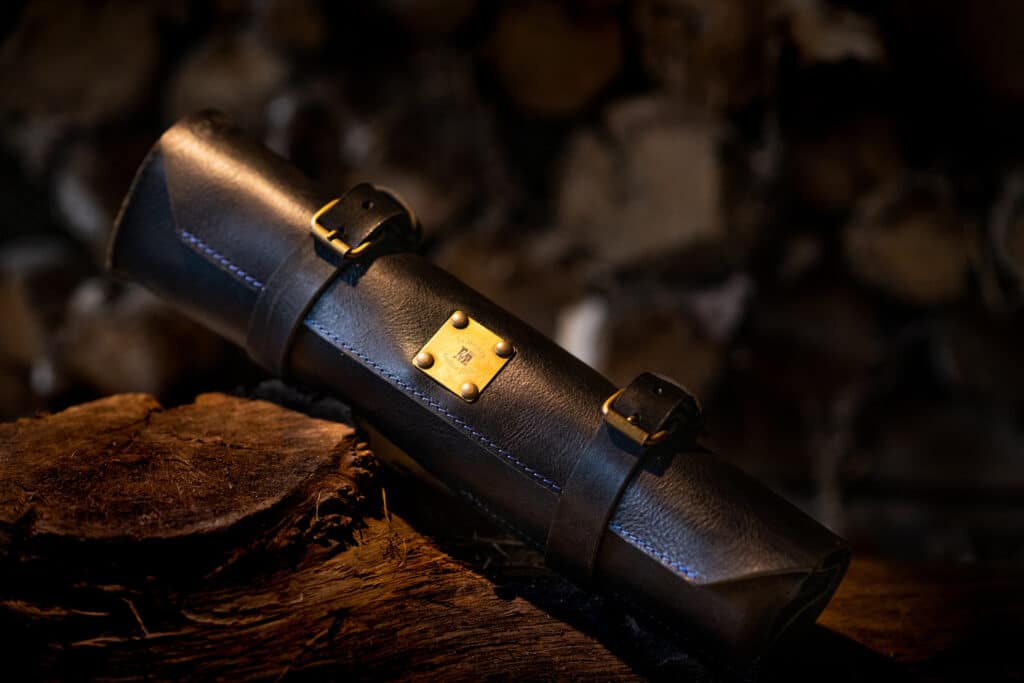 A cylindrical leather knife storage case secured by brass straps and nails on a rustic wood base.