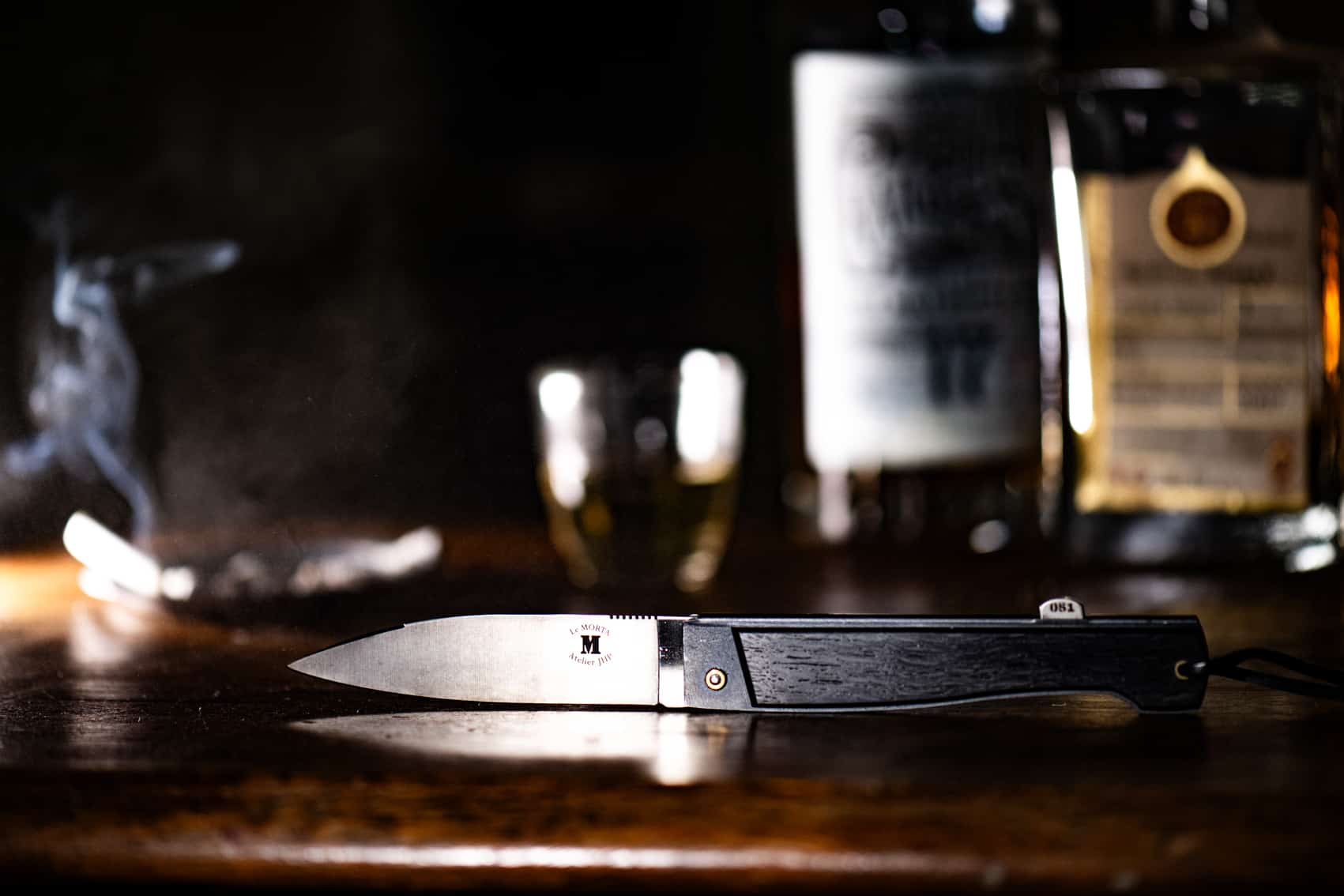 Un élégant couteau pliant avec un manche sombre et une lame en acier, reposant sur une surface en bois avec des volutes de fumée et des bouteilles en arrière-plan.