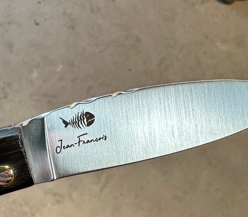 Lames d’émotions sur nos couteaux gravés pour l’éternité 2 Gros plan sur la lame d'un couteau, finement gravée du nom "Jean-Francois" et d'un pictogramme de poisson, incarnant une touche personnalisée des Couteaux Morta.
